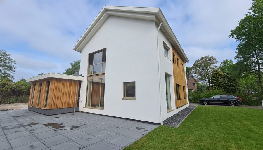 Zijgevel van de woning met een dubbelhoge puí en houten gevelbekleding, gezien vanaf de oprit
