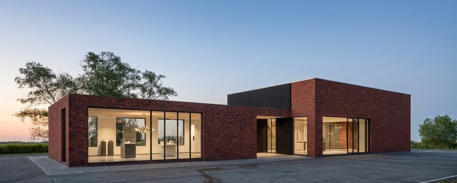 Ontwerp voor een lage showroom in donkere baksteen met een doorlopende glasstrook bij de entree, bij avondlicht