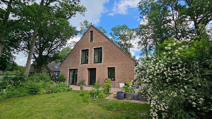 Bakstenen kopgevel van de woning met zadeldak, tussen volwassen bomen